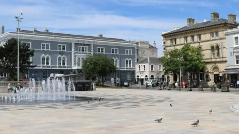 Google The Italian Gardens public plaza in Weston super Mare on a sunny day. It is a wide paved outdoor area with water fountains and lots of pigeons wandering around. It is surrounded by old pretty buildings and trees.