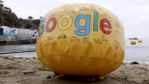 Google A Google-branded signed yellow buoy sits on the sand in as the first line is dropped