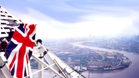 Tony Brien A photograph showing steel workers attaching the pyramid top to the skyscraper, with the River Thames and city behind.