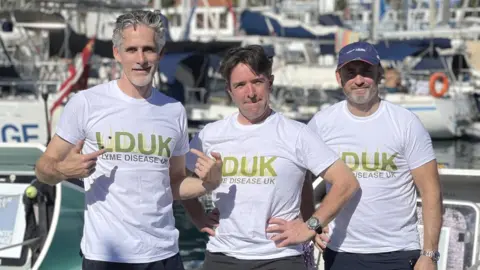 John Watling John Watling and two fellow rowers standing in a boat wearing Lyme disease T-shirts