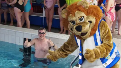 Lions Club of Jersey A man in a swimming pool wearing black swimming goggles and swimming shorts gives two thumbs up and smiles while waist deep in the water. A lion mascot wearing a large blue high-vis jacket is sat on the side of the pool also giving two thumbs up. A group of women are stood on the outside of the pool wearing swimming costumes.