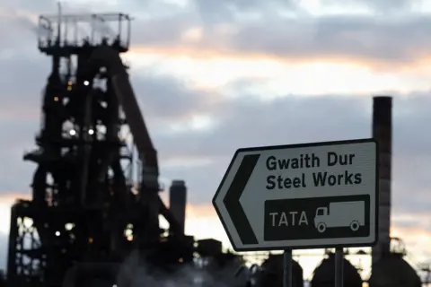 Reuters A road sign reads "Gwaith Dur Steel Works" and the company name Tata alongside a graphic of a lorry. In the background is one of the decommissioned blast furnaces at the Port Talbot works.