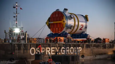 EDF A cylinder reactor on a huge boat. It dwarfs a boat next to it and containers underneath it. A concrete port is in the foreground with workers in visibility clothing dotted around.