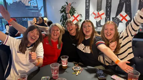 Five women at the England football watch party at Ashton Gate Stadium in Bristol. Three of them are in England football shirts. They all have their arms up in the air and are smiling at the camera.