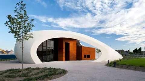 Philip Vile Maggie's in Aberdeen. It is a white building in the shape of a pebble or shell. It has a wooden entrances with grass and a tree in front of the building.