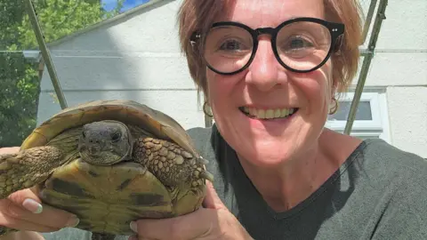 Fiona Sherriff A woman wearing round black glasses smiles as she holds up a tortoise to the camera.