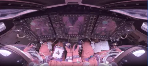 NASA This image looks forward over the shoulders of two Artemis II astronauts strapped into their seats inside the Orion crew module. They wear bulky orange pressure suits and clear helmet bubbles, facing a wall of tall, glowing display screens and dense rows of switches and knobs. Their gloved hands rest near hand controllers and a checklist. Around the edges of the frame, curved windows, cables and equipment make the cabin look compact and highly technical