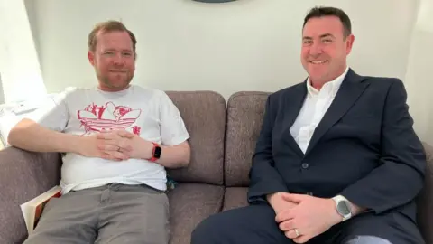 Two men sit on a sofa, smiling at the camera. One is wearing a T-shirt and shorts, the other a dark suit and white shirt.