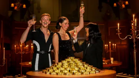 Studio Lambert/BBC/PA Stephen, Rachel and Claudia Winkleman hold flutes of champagne in front of a table full of golden pucks as they celebrate the show's final. Stephen is wearing a black and white striped jumpsuit with Rachel wearing a black sequined dress.