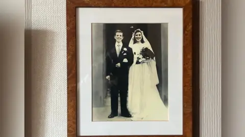 Adrian Harms/BBC A picture of Ted and Ellen on their wedding day. The pair, who are wearing a black suit and white dress respectively, are both smiling. The picture is in a brown frame. 