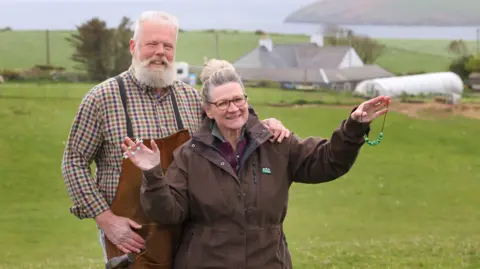 Colin Hattersley A man and a woman standing in a field near the Mull of Galloway. They are smiling at the camera and the woman is holding a beaded necklace in her hands. 