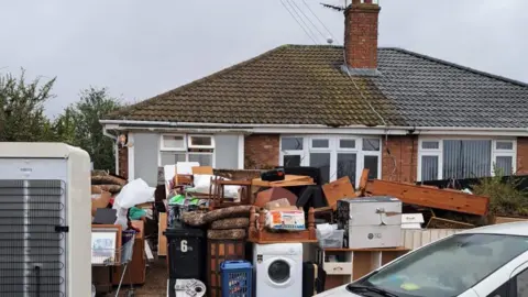 Submitted Piles of items including washing machines and furniture are piled up outside the property. They are stacked in the front garden, with the bungalow behind.