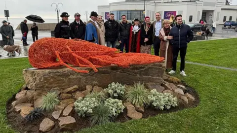 Bridlington ambassadors A group of officials stand behind the sculpture. A couple standing on the esplanade are standing beneath a black umbrella.
