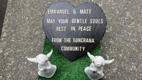 A heart shaped sign saying: 'Emmanuel & Matt May Your Gentle Souls Rest In Peace From The Buncrana Community' with two angel statues next to it.