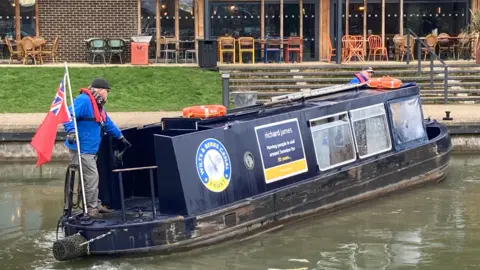 A man is driving a barge on a canal. The barge is blue. The man is driving it towards the towpath.