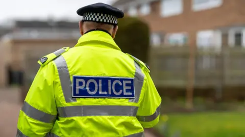 A police officer walks away from the camera, wearing a yellow high viz jacket with silver reflective stripes down the sides and a peaked hat with brown and white check around the rim. The background is blurred but shows a housing estate.