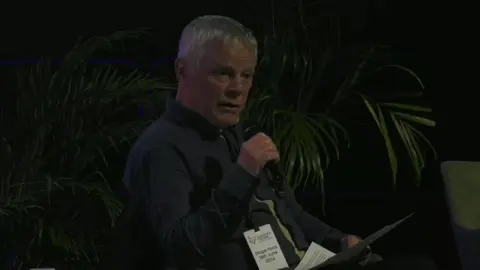 White haired man sitting and speaking at event