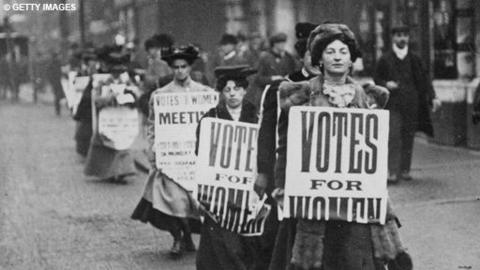 Suffragettes march in a line holding signs stating 'Votes for Women'.