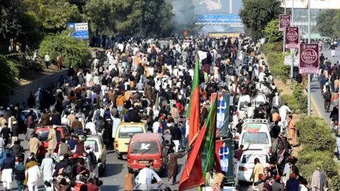 Reuters Hundreds of people walked along the main road lined with trees on both sides, with a few cars among the protesters. Some people are holding big red and green flags. Smoke can be seen rising in the distance.