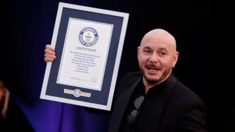 Pitbull wears a dark suit jacket over a dark shirt. He is holding a Guinness World Record award. 