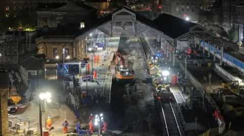 A railway station at night with several people dressed in orange on the platforms along with machinery. The platform is lit up by floodlights. 