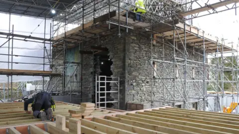 Freshfield Common barn during the restoration work. Scaffolding has been erected around the building. One workman stands on the scaffolding near the roof, while another is leaning on wooden beams on the first floor.