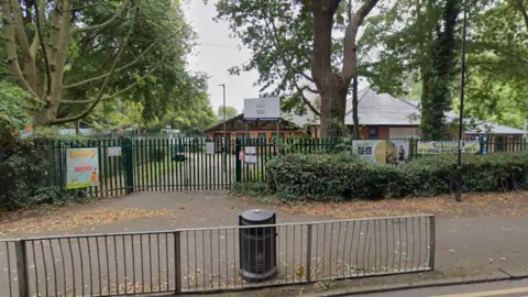 A gated entrance to a school with a low fence, signs on the gates, and buildings visible inside among the trees.