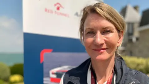Fran Collins is slim with dark blonde hair. She is wearing a red lanyard around her neck and a dark navy jacket over a shirt. The background is out of focus, but shows a banner reading 'Red Funnel', with bushes and the sea further in the background.