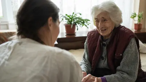 An older woman with grey hair smiles as she holds hands with a younger woman