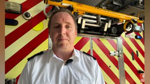 Zac Sherratt/BBC Dan Quin, chief fire officer for the Surrey Fire and Rescue Service. Dan is wearing a white shirt and is stood in front of a fire engine.