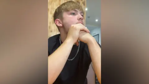 A man with blonde hair, in a black top with a silver chain necklace, looking away from the camera.