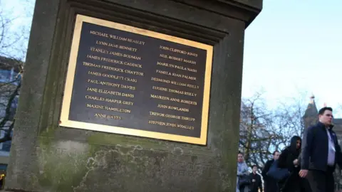 PA Media A stone memorial column with a large gold edged brown plaque showing 21 names written in gold. Pedestrians can be seen walking past