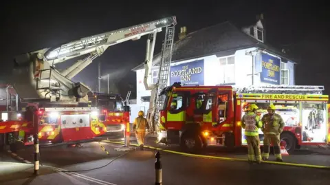 Fire appliances, including one with an extendable ladder, stand in front of the Portland Inn at Gurnard at night. The two-storey pub is illuminated by fire service lights. A handful of firefighters stand in the road.