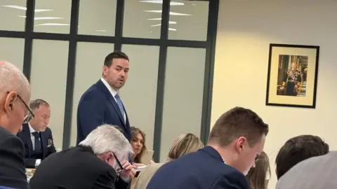 A man wearing a blue suit, standing up in a council chamber talking. There is a small portrait of King Charles III on the wall.