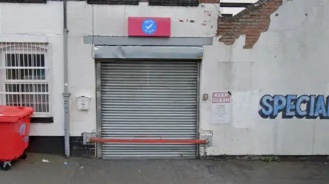 A grey shutter is down on a shop front that has a pink sign above it with a blue tick. The wall either side of the shutter is cream with graffiti on the right. 