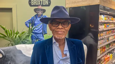 Wilfred Emmanuel-Jones is wearing a black glasses, a blue hat and a blue blazer, he stood infront of a model cow and drinks cabinet inside of his store 'The Black Farmer'.