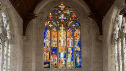 A view of the whole east window in an otherwise plain chapel with unadorned stone walls and a wooden roof.