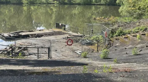 A stepped concrete bowl with broken wooden planks and a flat wooden area at the bottom, behind metal railings. There are weeds growing over the bowl. It faces a lake.