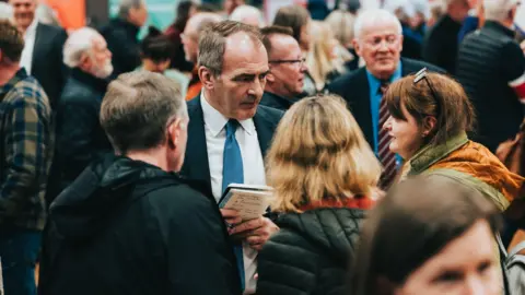 Isle of Man Government Alfred Cannan stands in the middle of a crowd of people, he talks to a woman wearing a green and yellow coat. He holds a pad and pen in his hands and wears a navy blue suit.