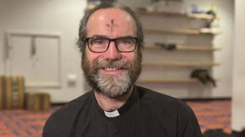 Head and shoulder shot of smiling middle-aged man. He has dark greying hair and beard, and is wearing spectacles. There is a faint cross marked in ash on his forehead. His black top and dog collar proclaim him as a chaplain.