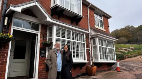 The pair are stood outside their pub in Clent. Chris has a long, beige jacket on with a blue shirt ad dark trousers on and brown shoes. He has a beard and grey hair. Lana is dressed all in black with white trainers and had long dark hair.