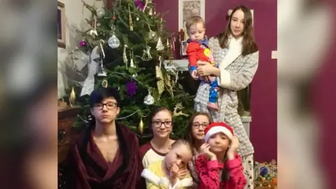 Family photo A group of seven children of various ages wearing pyjamas and dressing gowns in front of a Christmas tree. An older girl is standing holding a young child in her arms. The other five are sitting in front of her. The wall behind them is purple. 