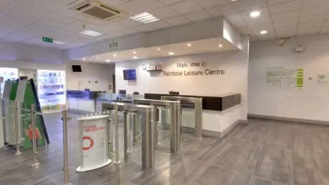 The interior of a leisure centre. There is a reception desk, turnstiles and a fridge.
