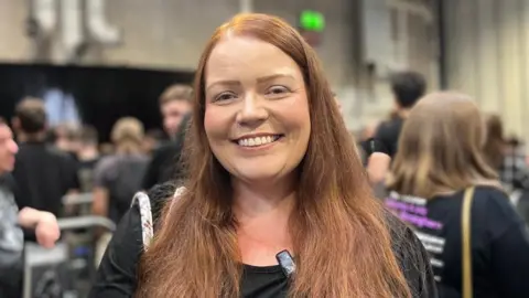 A woman with long hair is wearing a black top and smiling. She is standing in a queue with other people.
