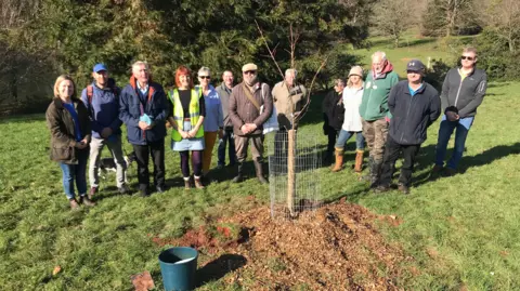 A group of volunteers stand in a line behind a recently-planted cherry tree.