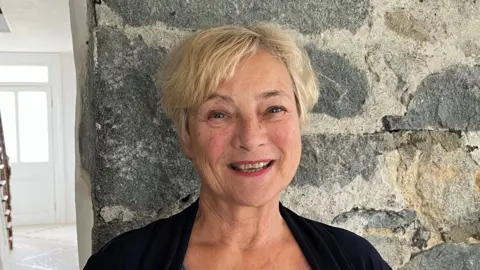 BBC A woman with short blonde hair, smiling at the camera, standing against a grey brick wall beside a hallway