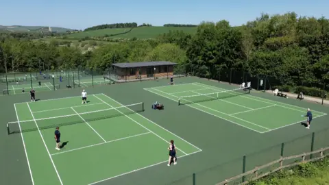 An aerial photo of Marlborough tennis club's six courts and new clubhouse. The courts have a green surface and there are several tennis matches taking place