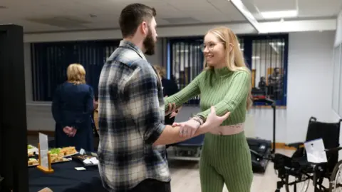 Chloe​​​​ O’Neill Megan being held up by Oliver, she is wearing a green top and green trousers, and a wheelchair is to get right. Oliver has his back to the camera and is wearing a checked top. Megan is smiling at him. 