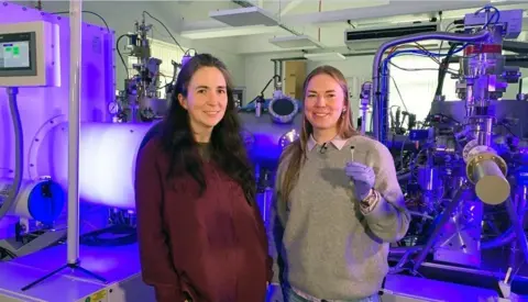 Loughborough University Prof Kelly Morrison (left) with Dr Naëmi Leo, in front of the Loughborough University nanolithography system. Dr Leo holds the chip that features the image of "the world’s smallest violin"