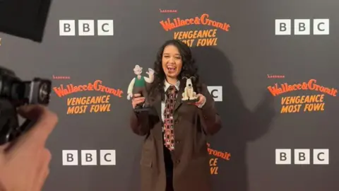 LaurenPatel Lauren Patel smiles holding a figurine in each hand of both Wallace and Gromit as a cameraman takes photos of her on the red carpet at the BAFTAs